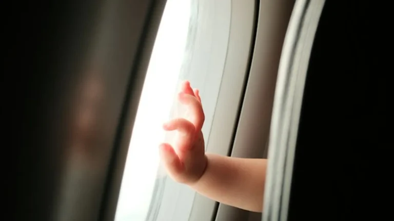 A baby's hand pressed against an airplane window in soft light — flying with a baby