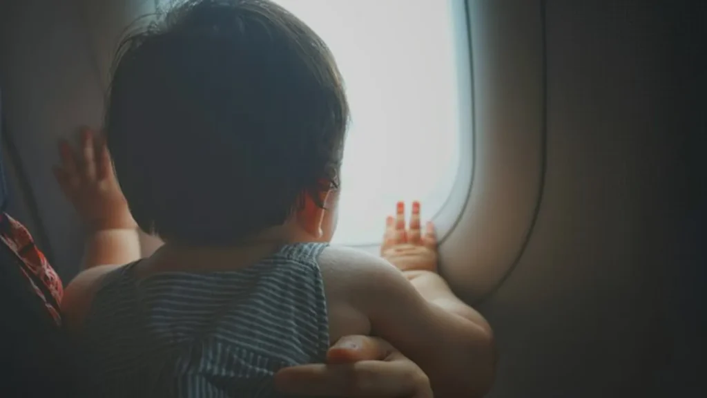 A toddler looking out of an airplane window, seen from behind, held by a parent's hand