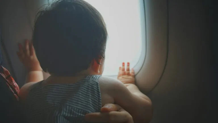 A toddler looking out of an airplane window, seen from behind, held by a parent's hand