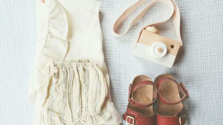 Minimal flat lay of baby clothes, tiny sandals and a toy camera on a cream muslin background